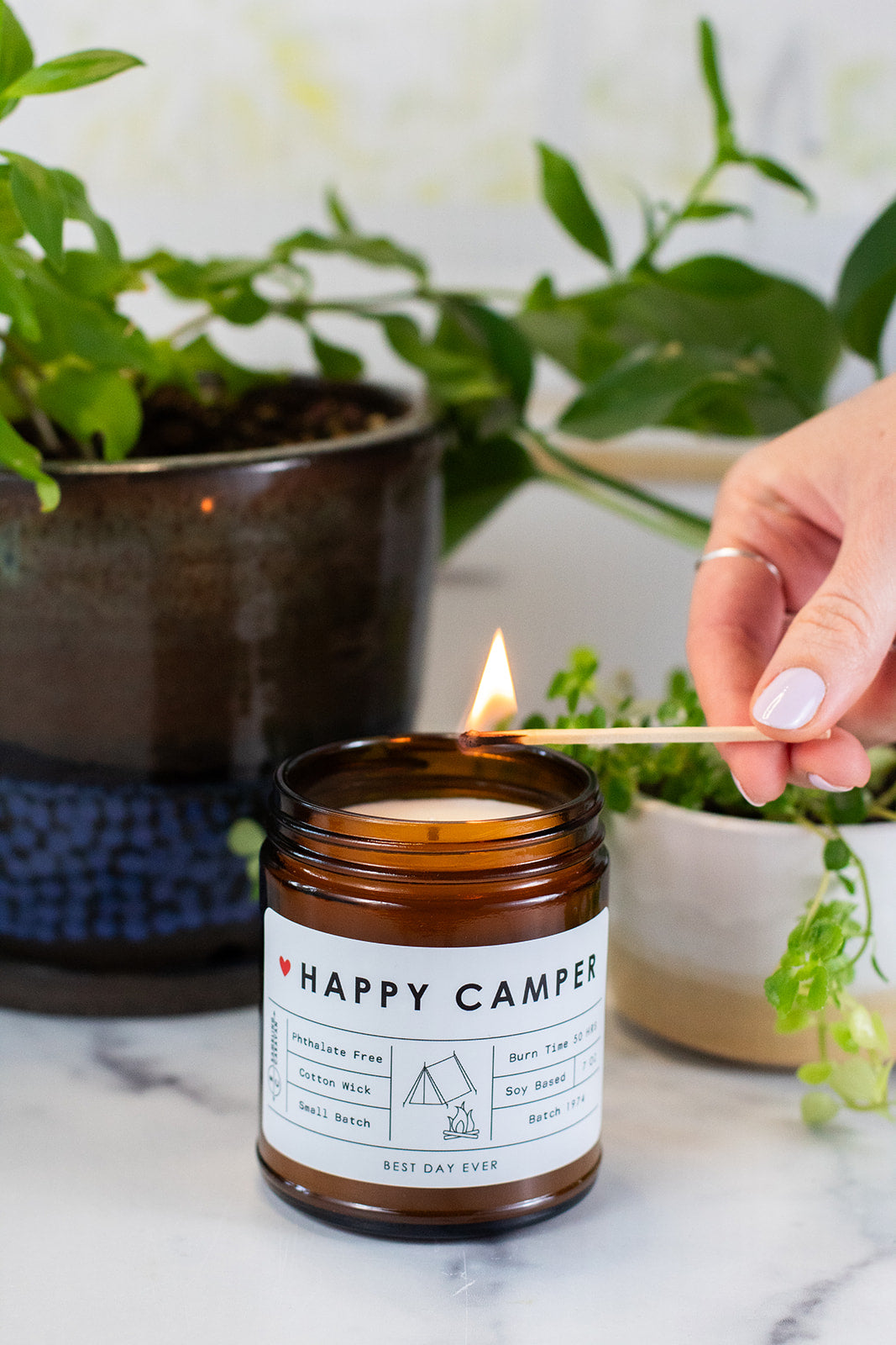 Happy Camper candle getting lit by match with plants in the background.